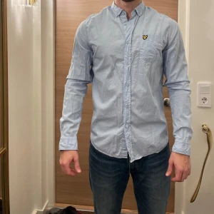 Ljusblå skjorta från Lyle & Scott - Snygg ljusblå skjorta från Lyle & Scott med klassisk krage och knappstängning. Skjortan har ett broderat emblem på bröstfickan och långa ärmar. Perfekt för en stilren look!