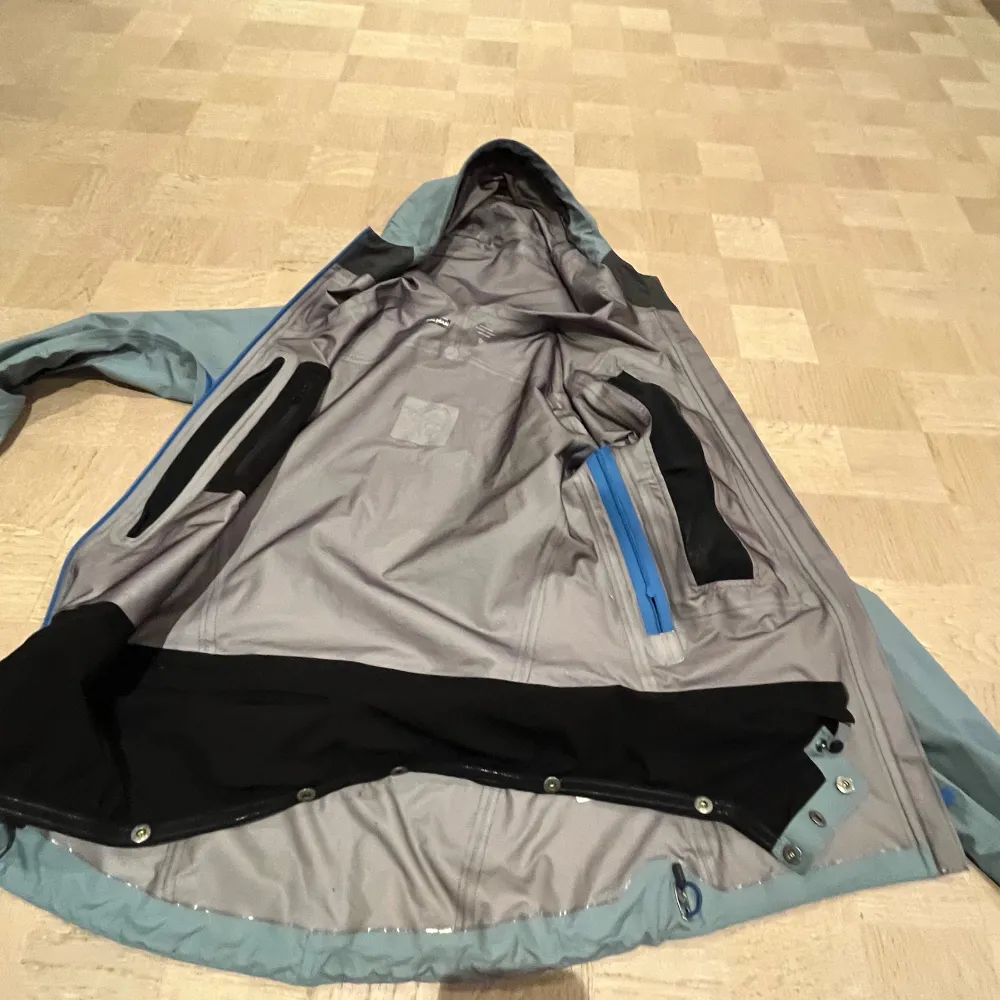 Säljer en grå Gore-Tex Lofoten jacka från Norrøna med huva och flera praktiska fickor med dragkedja. Jackan har en stilren design med blå detaljer och är perfekt för utomhusaktiviteter. Den är långärmad och tillverkad i ett slitstarkt material som skyddar mot väder och vind.. Takit.
