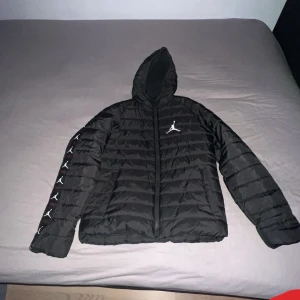 Svart pufferjacka från Jordan - Säljer en svart pufferjacka från Jordan med ikonisk logga på bröstet och ärmen. Jackan har en dragkedja framtill och en huva för extra skydd. Perfekt för kyliga dagar. 13-15 år, 163-175 cm