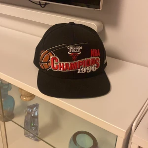 Svart keps från Mitchell & Ness  - Snygg svart keps med broderad text och motiv för Chicago Bulls NBA Champions 1996. Kepsen har en justerbar baksida och är perfekt för basketfans. Märket Mitchell & Ness är broderat på sidan. Det ända är det ni ster på sista bilden under kepsen. Det är några slags vita linjer men dom borde gå bort om man tar sig tiden!