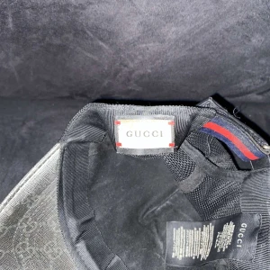 Svart keps med orm från Gucci - Snygg svart keps från Gucci med ett coolt ormmotiv i rött, vitt och blått. Kepsen har ett diskret mönster med Guccis logotyp och justerbart spänne baktill.