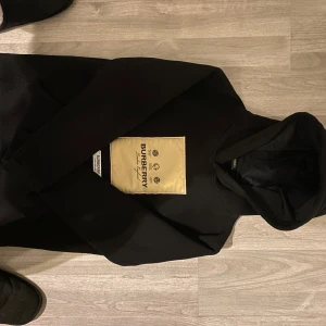 Svart hoodie från Burberry - Säljer en stilren svart hoodie från Burberry med en stor beige logga på framsidan. Tröjan har en klassisk huva och långa ärmar. Perfekt för en avslappnad och trendig look. Priset kan alltid diskuteras