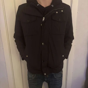 J lindeberg field jacket - Snygg svart jacka. Jackan har knappar och flera fickor framtill, vilket ger en praktisk och stilren look. Perfekt för kyligare dagar.