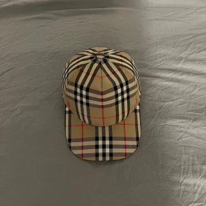 Rutig keps från Burberry - Snygg rutig keps från Burberry i klassiskt beige, svart och rött mönster. Kepsen har en justerbar rem baktill och är perfekt för en stilren look. Kommer med originalförpackning och påse. Och det är storlek L på den men samt passar M