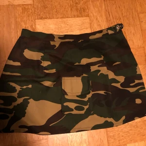 Camouflage kortkjol med dragkedja - Snygg kortkjol i camouflage-mönster med praktisk ficka framtill. Kjolen har en dragkedja och en praktisk ficka framtill 