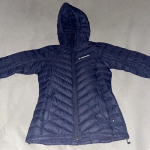 Blå dunjacka från Peak Performance - Säljer en snygg blå dunjacka från Peak Performance. Jackan har en praktisk huva och dragkedja framtill. Perfekt för kyliga dagar med sin quiltade design som håller värmen.