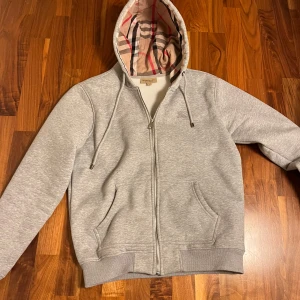 Grå hoodie från Burberry - Säljer en stilren grå hoodie från Burberry med klassiskt rutigt mönster i huvan. Tröjan har dragkedja och långa ärmar, perfekt för en avslappnad look. Passar utmärkt till både vardag och chill.