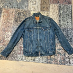 Jeansjacka från Levi's - Klassisk blå jeansjacka från Levi's med knappar framtill och två bröstfickor. Jackan har en tidlös design med en bekväm passform och är perfekt för både vår och höst. Den ikoniska röda Levi's-loggan syns på bröstfickan.