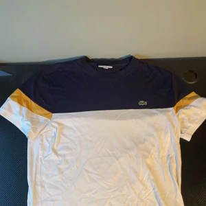 Lacoste tröja  - Säljer en snygg t-shirt från Lacoste i blått, vitt och gult. T-shirten har en klassisk design med korta ärmar och Lacostes ikoniska logga på bröstet. Perfekt för en avslappnad stil.