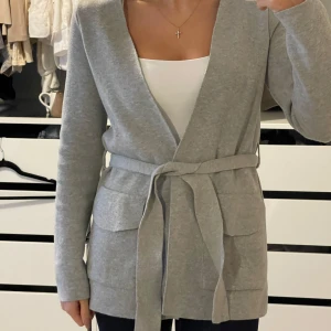 Grå kofta från Gina Tricot - Säljer en stilren grå kofta från Gina Tricot. Den har långa ärmar och ett knytbälte i midjan för en snygg passform. Perfekt för lager-på-lager-stil. Två praktiska fickor framtill. Passar till många olika outfits!