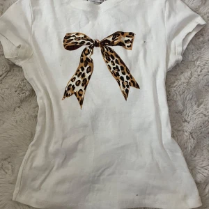 Vit t-shirt med leopardmönstrad rosett från Stradivarius - Säljer en vit t-shirt från Stradivarius med en stor leopardmönstrad rosett på framsidan. T-shirten har korta ärmar och en normal passform. Perfekt för att ge en touch av djurmönster till din outfit. 🐆