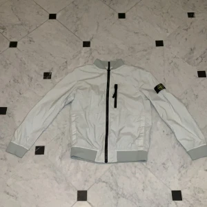 Vit bomberjacka från Stone Island Junior - Säljer en stilren vit bomberjacka från Stone Island Junior. Jackan har en svart dragkedja och en ikonisk logga på ärmen. Den är långärmad med ribbade muddar och krage. Perfekt för en cool och avslappnad look.
