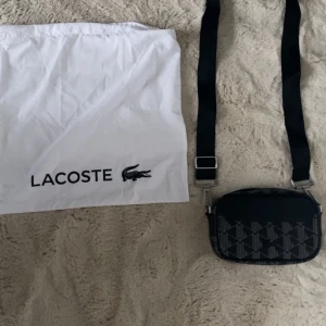 Lacoste axelväska - Snygg svart axelväska från Lacoste med ett geometriskt mönster i grått. Väskan har en justerbar axelrem och en dragkedja upptill. Perfekt för att bära dina nödvändigheter med stil. Inkluderar en vit dustbag med Lacoste-logga. Nypris på zalando är 1500kr