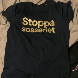 Svart t-shirt med tryck - Säljer en svart t-shirt med det guldiga trycket 'Stoppa sosseriet' på framsidan. T-shirten har en klassisk passform och är kortärmad, perfekt för en avslappnad stil.
