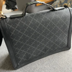 Svart handväska från By Malene Birger - Snygg svart handväska med ett diskret rutmönster från By Malene Birger. Väskan har en praktisk dragkedja och ett stilrent handtag. Perfekt för att bära med sig det viktigaste med stil. Hantag(rem) slitet men går att få fen utbytt. 