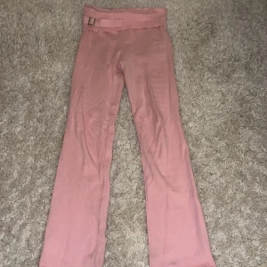 Rosa byxor med detalj - Snygga rosa byxor i 100% akryl med ett brett ribbat midjeband och dekorativt bälte. Byxorna har en rak passform och är perfekta för en avslappnad stil.