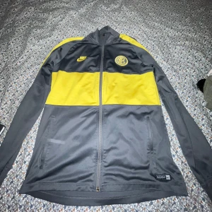 Inter Milan gul och grå fotbolls tröja  - Säljer en grå och gul träningsjacka från Nike med dragkedja framtill. Jackan har långa ärmar och är tillverkad i Dri-FIT-material för optimal komfort under träning. Perfekt för sportiga aktiviteter.