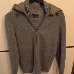 Stickad cardigan från Massimo Dutti - Säljer en stilren grå stickad tröja från Massimo Dutti. Tröjan har en praktisk dragkedja framtill och en avtagbar huva. Perfekt för kyligare dagar med sin mjuka och bekväma passform. 