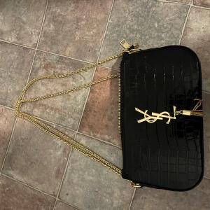 Svart axelväska från YSL - Snygg svart axelväska från YSL med krokodilpräglat mönster och guldfärgad kedja. Väskan har en elegant YSL-logga på framsidan och en praktisk dragkedja upptill. Perfekt för att ge en lyxig touch till din outfit.
