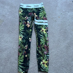 Mönstrade leggings från Stronger - Snygga leggings från Stronger med tropiskt blommönster i grönt, rosa och gult. Dem är ganska använda och har lite nåpror ner till med vaden, men annars bra skick 