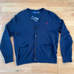 Mörkblå kofta från Ralph Lauren - Säljer en stilren mörkblå kofta från Ralph Lauren med v-ringning och knappar framtill. Den har två praktiska fickor och det ikoniska röda logotypbroderiet på bröstet. Perfekt för en klassisk look. Nypris: 2100 kr 