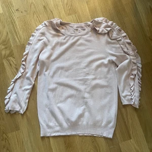 Ljusrosa tröja med volangdetaljer från Philosophy - Säljer en ljusrosa tröja från Philosophy med volangdetaljer längs ärmarna. Tröjan har trekvartsärm och är tillverkad i ett mjukt material. Perfekt för en stilren och feminin look.