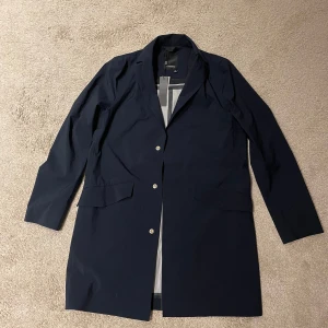 Mörkblå jacka från J.Lindeberg - Snygg mörkblå rock/windbreaker från J.Lindeberg med klassisk design och knappar framtill. Jackan/windbreakern är helt ny emn säljs pågrund av fel storlek. Jackan/windbreakern har långa ärmar och är perfekt för säsongens kyligare dagar. Den är tillverkad i ett slitstarkt material och har en elegant insida. Jackan/windbreakern är i storlek 46. Tips! Den passar bra om du är 180–185cm och kan vara lite för stor för dig som är 170-177 cm. Pris kan diskuteras.  Skriv vid det minsta lilla!🧥