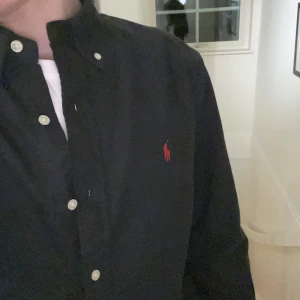 Svart skjorta från Ralph Lauren - Snygg svart skjorta från Ralph Lauren med klassisk krage och knappar. Den har en röd broderad logga på bröstet som ger en stilren touch. Perfekt för en elegant look.