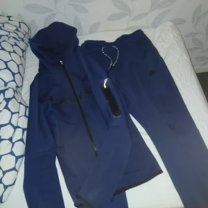 Mörkblå nike tech tracksuit dress - Säljer en blå hoodie och matchande byxor från Nike. Hoodien har dragkedja och en stilren design med svarta detaljer. Byxorna har dragsko i midjan för justerbar passform. Perfekt för en avslappnad stil. Inga fläckar eller hål nästan som ny. Tröjan s men sitter som M byxorna M sitter som M
