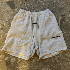 Grå shorts från Essentials - Säljer ett par grå shorts från Essentials med elastisk midja och snörning. Perfekta för en avslappnad stil. Essentials-loggan är tryckt på framsidan. Ej äkta