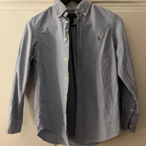 Blå skjorta från Ralph Lauren - Snygg blå skjorta från Ralph Lauren med klassisk krage och knappar framtill. Skjortan har långa ärmar och en broderad logotyp på bröstet. Perfekt för en stilren look.  Helt o använd
