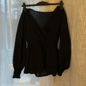 Svart omlottblus - Snygg svart omlottblus från Zara med långa ärmar och smockdetaljer i midjan. Blusen har en v-ringad design och är perfekt för en stilren look. Materialet är lätt och bekvämt.