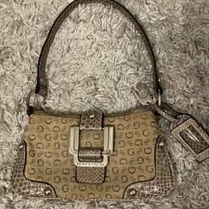 Beige axelväska från Guess - Snygg beige axelväska från Guess med mönstrat tyg och ormskinnsdetaljer. Väskan har en praktisk dragkedja och ett dekorativt spänne framtill. Perfekt för att bära dina nödvändigheter med stil. 