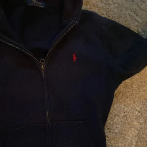 Mörkblå hoodie från Ralph Lauren - Säljer en snygg mörkblå hoodie från Ralph Lauren med dragkedja och det klassiska röda logotypbroderiet på bröstet. Perfekt för en avslappnad stil. Den har en praktisk huva och långa ärmar.
