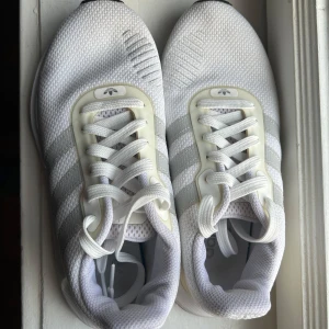 Vita sneakers från Adidas - Snygga vita sneakers från Adidas med klassiska tre ränder på sidan. Skorna har en stilren design med snörning och en bekväm sula i svart. Perfekta för en sportig look. Strl 39