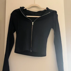 Svart ribbad hoodie med dragkedja - Säljer en svart ribbad hoodie med dragkedja framtill. Tröjan har långa ärmar och en croppad passform, perfekt för en avslappnad stil. Den är både stilren och bekväm.