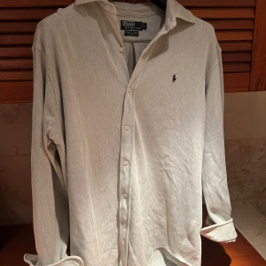 Beige skjorta från Polo Ralph Lauren - Snygg beige skjorta från Polo Ralph Lauren i 100% bomull. Skjortan har långa ärmar och knappar framtill. Den klassiska designen är perfekt för en stilren look. Broderad logotyp på bröstet ger en extra touch av elegans. Ganska stor i storlek sitter typ XL även om lappen säger L.