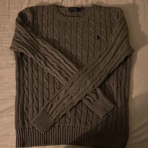 Grå stickad tröja från Ralph Lauren - Säljer en grå stickad tröja från Ralph Lauren med kabelstickat mönster. Tröjan har långa ärmar och en rund halsringning. Perfekt för kyligare dagar.