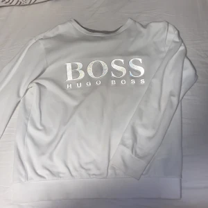 Vit tröja från Hugo Boss - Säljer en stilren vit tröja från Hugo Boss med deras logga i silver på bröstet. Tröjan har långa ärmar och en klassisk passform. Perfekt för en avslappnad och trendig look.
