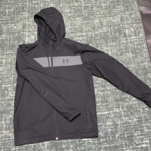 Svart vindjacka från Under Armour - Snygg svart vindjacka från Under Armour med huva och dragkedja. Jackan har en grå detalj över bröstet med logotyp. Perfekt för blåsiga dagar och sportiga aktiviteter.