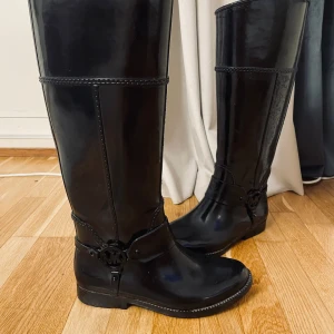 Svarta stövlar från Michael Kors - Snygga svarta stövlar från Michael Kors i syntetmaterial. De har en dekorativ rem med logotypdetalj runt ankeln och en klassisk hög design. Perfekta för en stilren look.