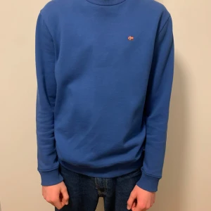 Blå tröja från Napapijri - Säljer en stilren blå tröja från Napapijri med en liten broderad logga på bröstet. Tröjan har långa ärmar och ribbade muddar vid ärmslut och nederkant. Perfekt för en avslappnad look.