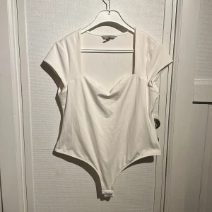 Vit bodysuit med korta ärmar - Snygg vit bodysuit med korta ärmar och en elegant skärning vid halsen. Perfekt för att skapa en stilren look. Tillverkad i ett mjukt och bekvämt material.