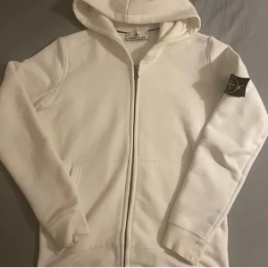 Vit hoodie från Stone Island - Säljer en stilren vit hoodie från Stone Island med dragkedja och huva. Den har en broderad logga på ärmen och är perfekt för en avslappnad look. Hoodien har långa ärmar och praktiska fickor framtill.