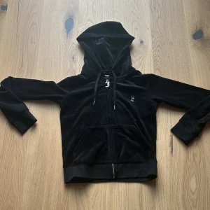 Svart velourjacka från Juicy Couture - Säljer en snygg och bekväm svart velourjacka från Juicy Couture. Den har en dragkedja framtill och en stor huva. På baksidan finns det ett glittrigt 'Juicy'-tryck. Perfekt för en avslappnad och stilren look!