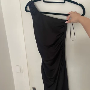 Svart oneshoulder klänning - Säljer en elegant svart oneshoulder klänning. Den har en tight passform och är perfekt för festliga tillfällen. Materialet är mjukt och följsamt, vilket ger en smickrande siluett. Klänningen har en stilren design med rynkade detaljer längs sidan den är även lång. Storlek XXS. Helt ny!