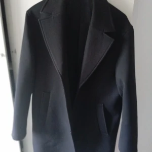 Svart kappa - Säljer en stilren svart kappa i storlek M. Den har en klassisk design med långa ärmar och två fickor framtill. Perfekt för höst och vinter för en elegant look.