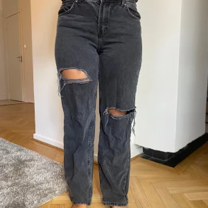 Svarta jeans med slitningar - Säljer ett par svarta jeans med coola slitningar på knäna. De är högmidjade och har en straight passform som ger en avslappnad look. Perfekta för en casual outfit eller när du vill lägga till lite edge till din stil.