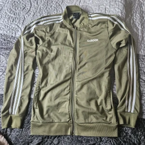 Grön Adidas jacka - Säljer en grön Adidas jacka i storlek S. Den har de klassiska tre ränderna längs ärmarna och en dragkedja framtill. Jackan är långärmad och i bra skick, men har ett litet hål på framsidan. Perfekt för träning eller vardagsbruk!