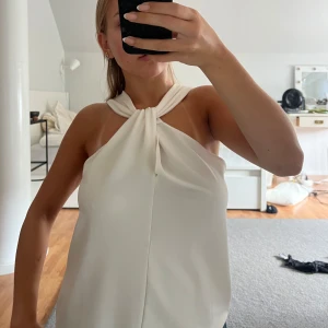 Vit axelbandslös topp - Säljer en stilren vit axelbandslös topp med en elegant knutdetalj vid halsen. Perfekt för sommarens fester eller en avslappnad dag ute. Toppen är i ett mjukt och bekvämt material som faller snyggt. Den är i nyskick å har aldrig använts!!